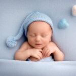 Neugeborener Junge schläft in den ersten Lebenstagen nackt auf blauem Stoffhintergrund in einer blauen Wollmütze.