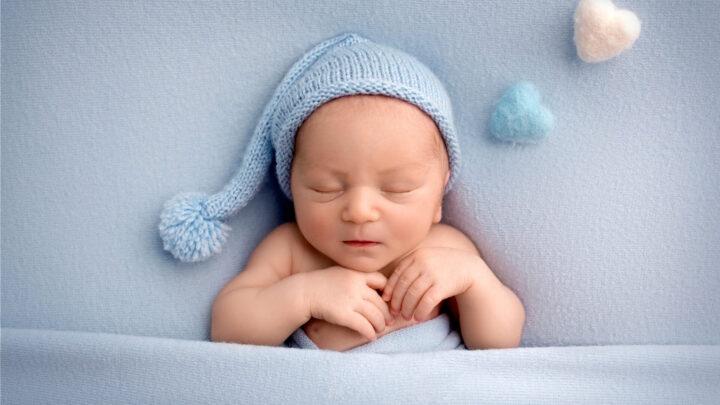 Neugeborener Junge schläft in den ersten Lebenstagen nackt auf blauem Stoffhintergrund in einer blauen Wollmütze.